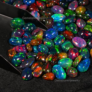May include: A collection of loose, colorful, iridescent opal gemstones in various shapes and sizes. The gemstones are displayed in a black metal scoop on a black surface. The text "Gemstonejewelry" appears twice in the image.
