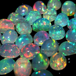 Puede incluir: Una colección de piedras preciosas de ópalo ovaladas e iridiscentes con una variedad de colores, incluyendo verde, azul, rojo y naranja. Las piedras preciosas están dispuestas en un patrón aleatorio sobre un fondo negro.