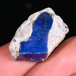Könnte beinhalten: Ein roher, ungeschliffener Opal-Edelstein mit leuchtenden blauen und grünen Farben. Der Opal ist in einer hellgrauen Gesteinsmatrix eingebettet. Der Edelstein wird in einer Hand gehalten.