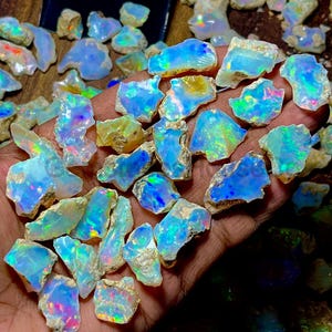 Puede incluir: Una colección de piedras preciosas de ópalo en bruto, que muestra un espectro de colores que incluye azules, verdes y destellos de tonos iridiscentes. Los ópalos tienen una textura rugosa y natural y tamaños variables, con una matriz de color claro.