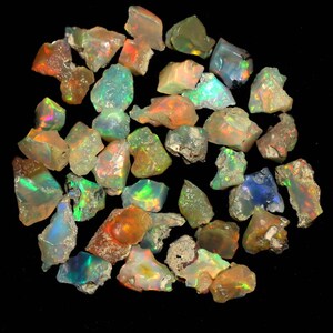 Puede incluir: Una colección de piedras preciosas de ópalo en bruto en varios tonos de verde, azul, naranja y blanco. Los ópalos son iridiscentes y tienen un acabado natural sin pulir.