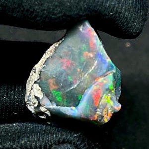 Puede incluir: Una piedra preciosa de ópalo negro en bruto con destellos vibrantes de rojo, verde y azul. El ópalo tiene un color base oscuro y una superficie texturizada e irregular. Una pieza única de joyería natural.