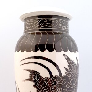 May include: A white ceramic vase with a brown dragon design. The dragon is detailed with intricate scales and has a fierce expression. The vase has a brown rim and a brown base.