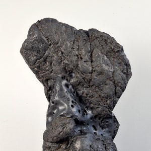 May include: A black volcanic rock sculpture with a rough, textured surface. The rock is displayed on a dark wood base.