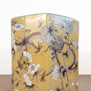 May include: A yellow ceramic square stool with a dragon and floral design. The dragon is black and gray and the flowers are white.