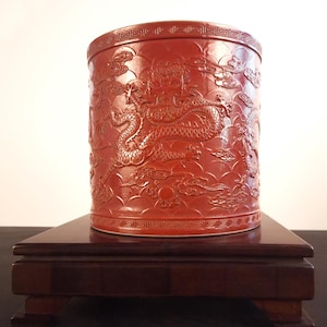 May include: A cylindrical, red ceramic container with a detailed relief carving of a dragon and clouds. The container sits on a dark wooden stand. The design features a repeating geometric pattern around the rim.