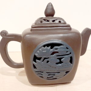 May include: A brown ceramic teapot with a square body and a lid with a small knob on top. The teapot has a carved design of a lotus flower and other floral elements on the side.