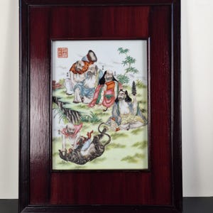 May include: A framed porcelain plaque featuring a colourful painting of five figures in a landscape setting. The figures are depicted in traditional Chinese clothing and are surrounded by bamboo plants and a mountain. The plaque is framed in a dark wood frame with a brass hanging loop.
