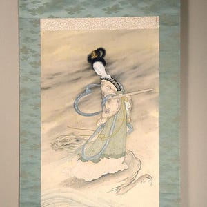 May include: A traditional Japanese scroll painting depicting a woman in a flowing robe standing on a wave. The painting is done in shades of blue, green, and white, with gold accents. The woman's hair is black and she has a serene expression on her face.