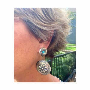 May include: Silver-toned, dangling earrings. The top portion is a small, round stud with a turquoise stone in the center. The bottom portion is an oval shape with a floral design and etched details. The earrings are worn on a person.