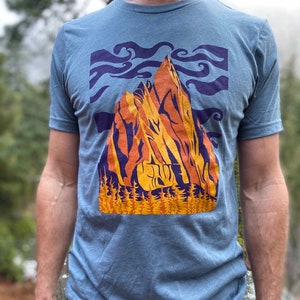May include: A blue t-shirt with a graphic of a mountain range in orange, yellow, and purple. The mountain range is surrounded by a blue sky with white clouds. The graphic is set against a blue background.
