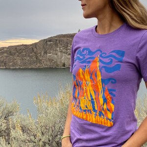 May include: A purple t-shirt with a graphic design of a mountain range with orange, yellow, and blue colors. The design includes a stylized depiction of trees and clouds.