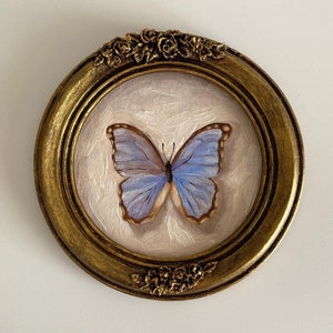 Puede incluir: Una pintura enmarcada de una mariposa azul con venas marrones en sus alas. El marco es dorado y tiene un diseño floral decorativo.