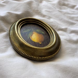 May include: An oval, antique gold-coloured frame featuring a painting of a yellow lemon and green leaves. The frame has a beaded edge and is set against a white, textured background. The artwork is a still life.