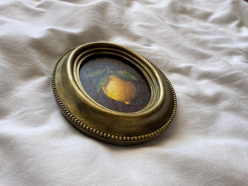 May include: An oval, antique gold-coloured frame featuring a painting of a yellow lemon and green leaves. The frame has a beaded edge and is set against a white, textured background. The artwork is a still life.