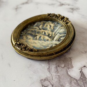 May include: A small oval-shaped framed painting of a winter scene with trees and snow. The frame is gold with a decorative design.