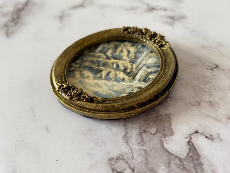 May include: A small oval-shaped framed painting of a winter scene with trees and snow. The frame is gold with a decorative design.