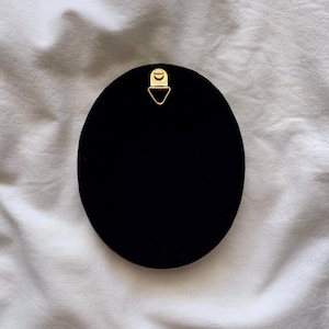May include: A black, oval object with a gold-coloured metal hanger at the top. The object is set against a white, textured background. The hanger is a small, triangular shape.