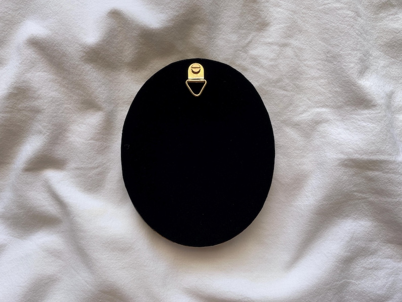 May include: A black, oval object with a gold-coloured metal hanger at the top. The object is set against a white, textured background. The hanger is a small, triangular shape.
