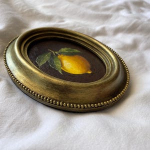 May include: Oval framed painting of a lemon with green leaves. The artwork is set against a dark background and framed in a gold-toned, ornate frame. The frame has a beaded edge and a slightly distressed finish.