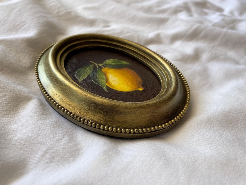 May include: Oval framed painting of a lemon with green leaves. The artwork is set against a dark background and framed in a gold-toned, ornate frame. The frame has a beaded edge and a slightly distressed finish.