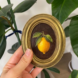 May include: Oval painting in a gold frame, depicting a lemon with green leaves on a dark brown backdrop. The painting is held in a hand, with a green plant visible. The frame has a detailed, textured edge.