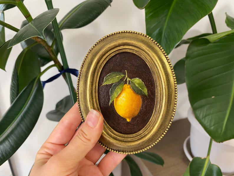 May include: Oval painting in a gold frame, depicting a lemon with green leaves on a dark brown backdrop. The painting is held in a hand, with a green plant visible. The frame has a detailed, textured edge.