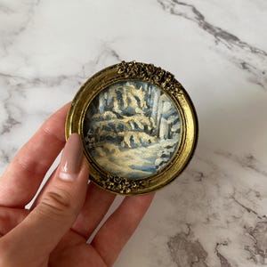 May include: A small, round, gold-framed picture with a winter scene painted inside. The scene depicts a snowy forest with trees and a path.