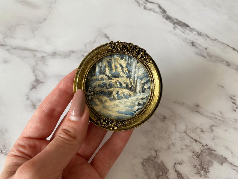 May include: A small, round, gold-framed picture with a winter scene painted inside. The scene depicts a snowy forest with trees and a path.