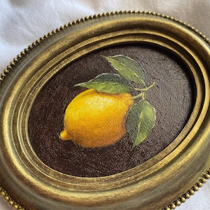 May include: Oval framed painting of a lemon with green leaves. The lemon is yellow with a slight orange hue, and the leaves are a vibrant green. The frame is gold with a beaded edge, set against a dark brown background.