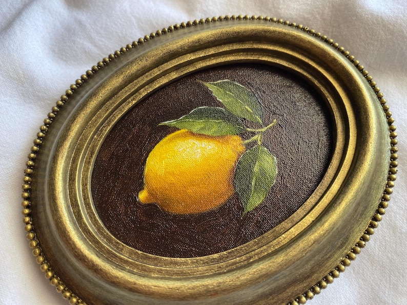 May include: Oval framed painting of a lemon with green leaves. The lemon is yellow with a slight orange hue, and the leaves are a vibrant green. The frame is gold with a beaded edge, set against a dark brown background.