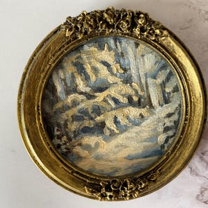May include: A small, framed painting depicting a snowy forest scene with a blue and white color scheme. The frame is gold with a decorative floral design.