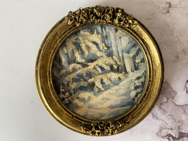 May include: A small, framed painting depicting a snowy forest scene with a blue and white color scheme. The frame is gold with a decorative floral design.