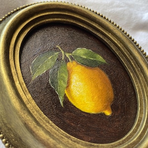 May include: Oval framed painting of a lemon with green leaves. The lemon is yellow with a brown background. The frame is gold with a beaded edge. The painting is a still life.