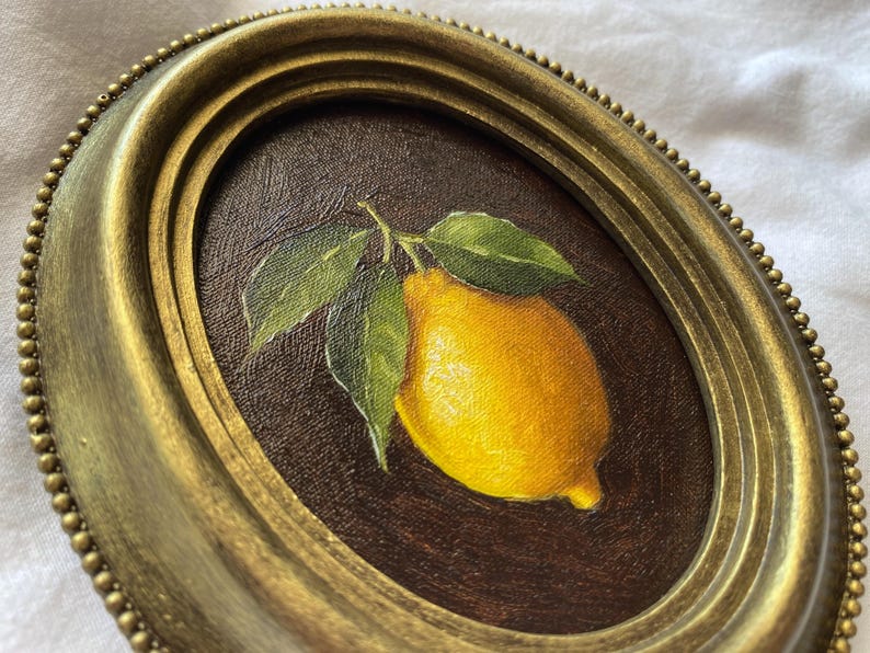 May include: Oval framed painting of a lemon with green leaves. The lemon is yellow with a brown background. The frame is gold with a beaded edge. The painting is a still life.