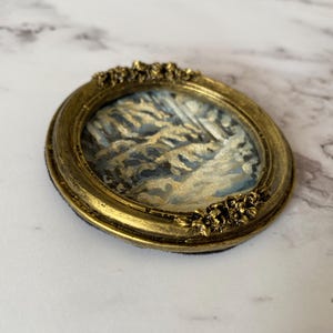 May include: A small, oval-shaped painting of a winter scene with a gold-colored frame. The painting depicts a snowy landscape with trees and a blue sky.