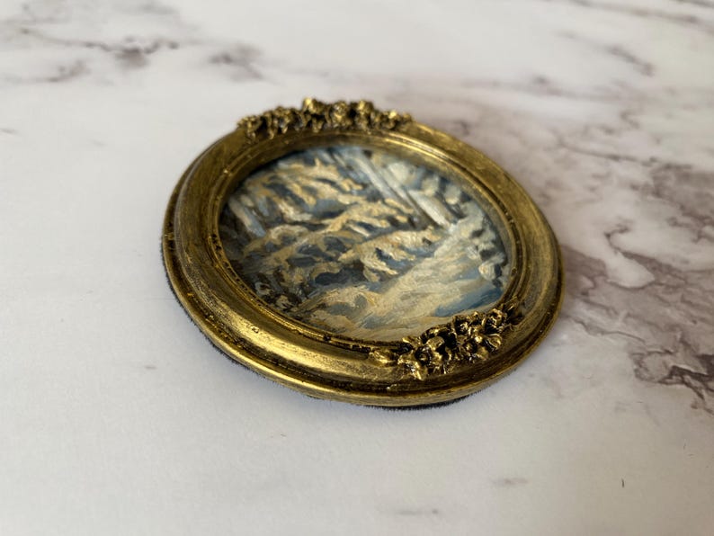 May include: A small, oval-shaped painting of a winter scene with a gold-colored frame. The painting depicts a snowy landscape with trees and a blue sky.