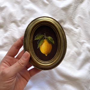 May include: Oval framed painting of a lemon with green leaves. The lemon is yellow, set against a dark brown background. The frame is gold-toned with a beaded edge. The painting is held against a white background.