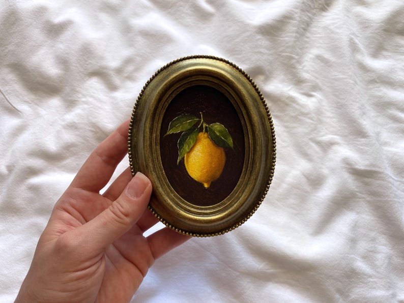 May include: Oval framed painting of a lemon with green leaves. The lemon is yellow, set against a dark brown background. The frame is gold-toned with a beaded edge. The painting is held against a white background.