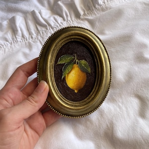May include: Oval framed artwork in an antique style, depicting a bright yellow lemon with green leaves on a dark brown backdrop. The frame is gold-toned with a beaded edge. The artwork is held by a hand.
