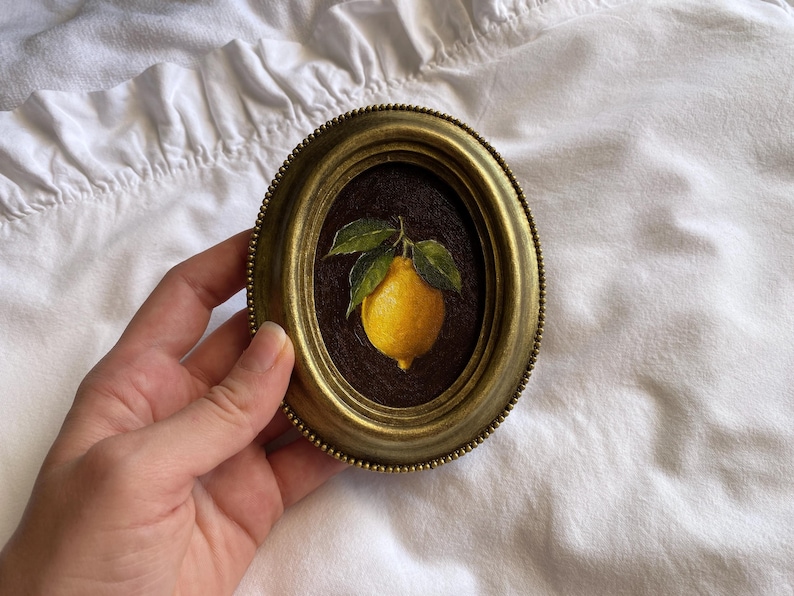 May include: Oval framed artwork in an antique style, depicting a bright yellow lemon with green leaves on a dark brown backdrop. The frame is gold-toned with a beaded edge. The artwork is held by a hand.