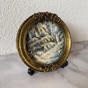 May include: A small, framed painting of a snowy winter scene with trees and a path. The frame is gold with ornate details and is on a black stand.