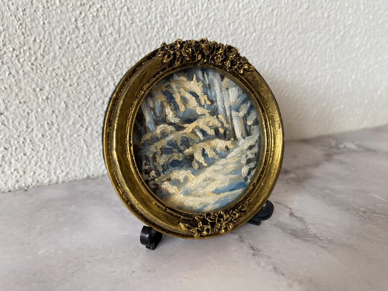 May include: A small, framed painting of a snowy winter scene with trees and a path. The frame is gold with ornate details and is on a black stand.