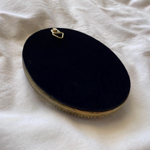 May include: An oval-shaped object with a black velvet surface and a gold-coloured, beaded trim. A small, gold-coloured metal hanger is attached to the top. The object is set against a white, textured background.