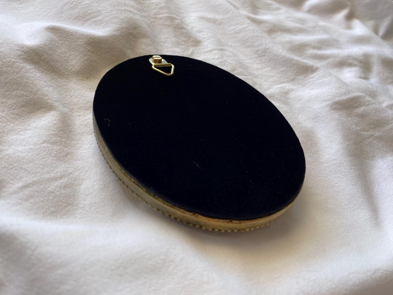 May include: An oval-shaped object with a black velvet surface and a gold-coloured, beaded trim. A small, gold-coloured metal hanger is attached to the top. The object is set against a white, textured background.