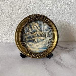 May include: A small, round, gold-colored frame with a winter scene painting inside. The painting depicts a snowy forest with trees and a path.