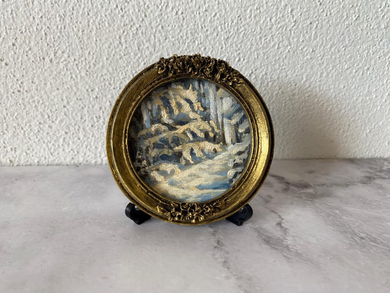 May include: A small, round, gold-colored frame with a winter scene painting inside. The painting depicts a snowy forest with trees and a path.