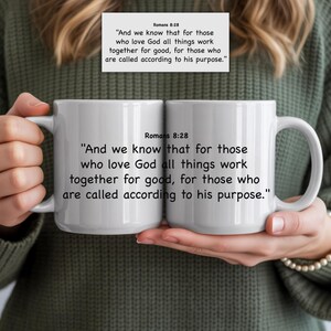 May include: Two white ceramic mugs with the text "Romans 8:28" and a Bible verse printed in black. The mugs are held by a person wearing a green jumper. The mugs are a standard size.