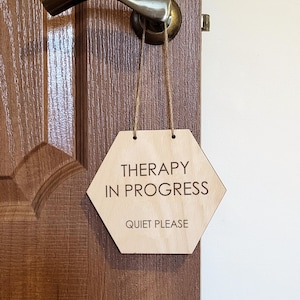 Könnte beinhalten: Ein sechseckiges Holzschild mit der Aufschrift "THERAPY IN PROGRESS QUIET PLEASE" hängt an einem Türgriff. Das Schild ist mit Schnur befestigt und aus hellem Holz gefertigt. Die Tür ist braun mit einem messingfarbenen Griff.