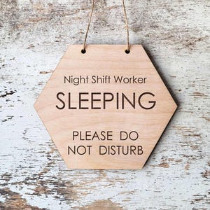 May include: A hexagonal light-colored wooden sign with a thin string for hanging. The text engraved in black reads: Night Shift Worker, SLEEPING, PLEASE DO NOT DISTURB. The sign features a smooth, natural wood grain finish and clean, sans-serif lettering.
